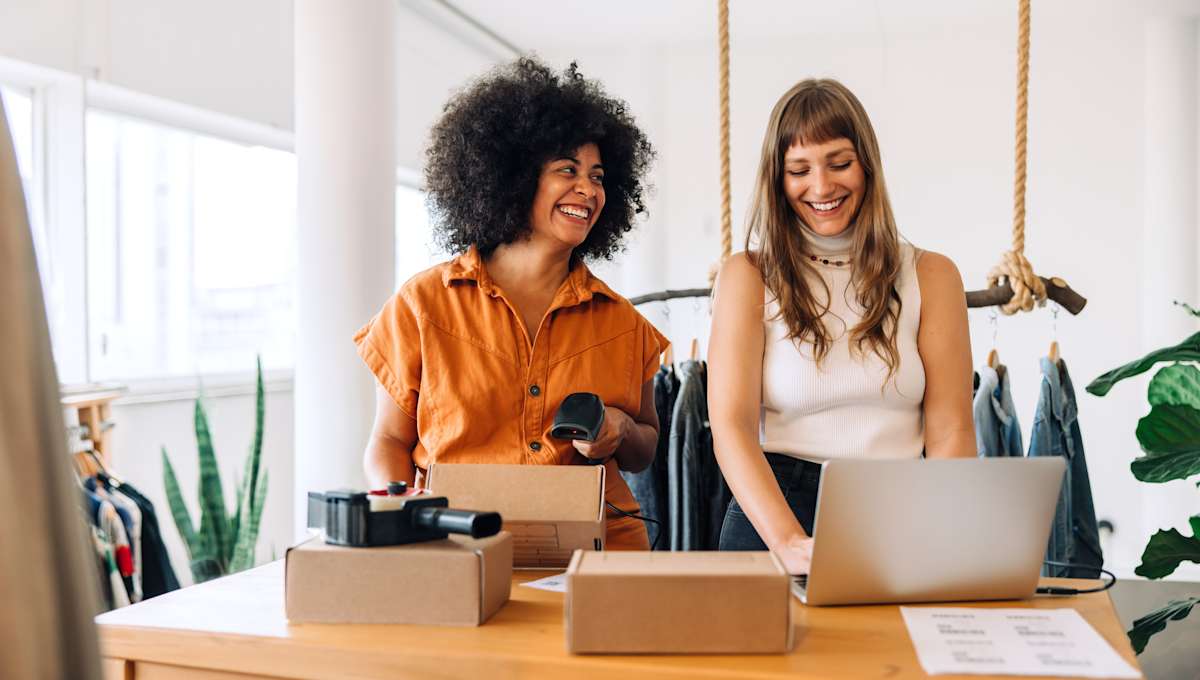 two women work on a laptop together with some packing boxes on a desk