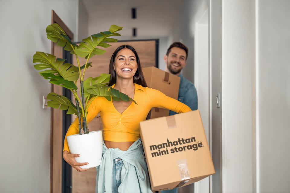 A Woman carries a plant and a Manhattan Mini Storage Moving box, while a man carries more moving boxes behind her