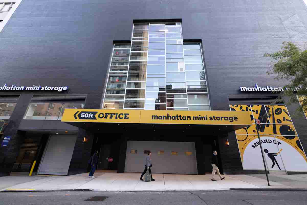 Street-level view of Manhattan Mini Storage at 420 E 62nd St in New York City, featuring a modern black building facade, large windows, bold yellow signage, and pedestrians walking by.