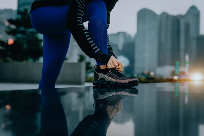 Confidence and energetic sports woman tying shoe laces and getting ready to exercise in urban park against city skyline at sunset