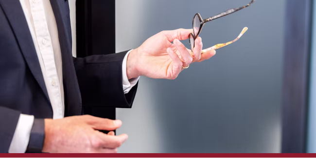 Man in suit gesturing and holding eyeglasses in his left hand.