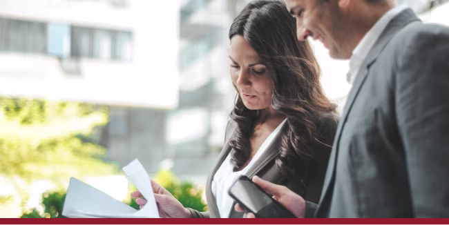Two business professionals engaged in a serious discussion while reviewing documents and using a smartphone in an outdoor urban setting.