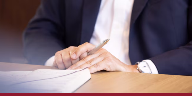 Men in a suit with watch on his wrist and a pen between his hands. He is sitting at a table with a notebook in front of him. 
