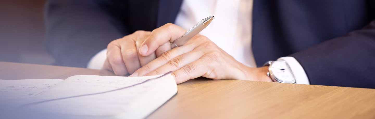 Men in a suit with watch on his wrist and a pen between his hands. He is sitting at a table with a notebook in front of him. 