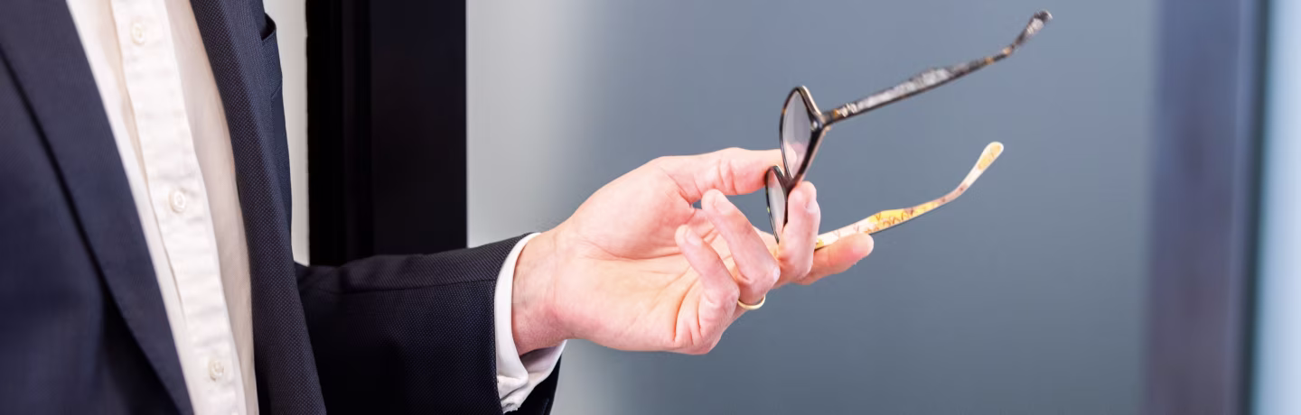 Man in suit gesturing and holding eyeglasses in his left hand.