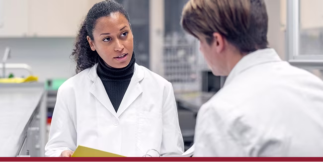 Female researcher holding a document file and discussing latest findings with male colleague in scientific lab.