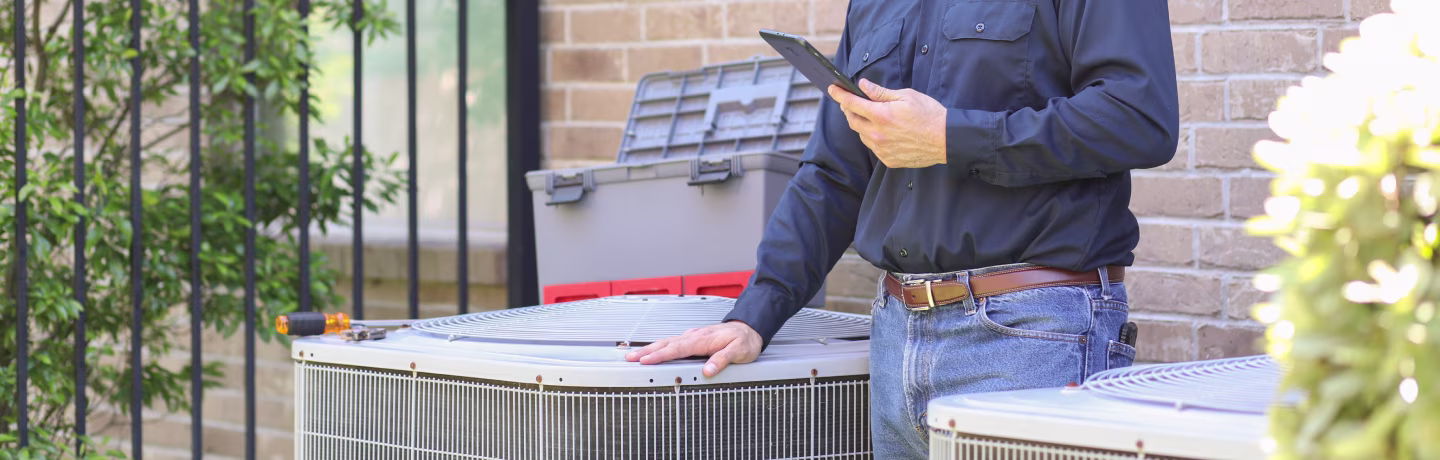 Blue collar air conditioner repairman working at residential home. He prepares to begin work by gathering appropriate tools and referring to digital tablet.