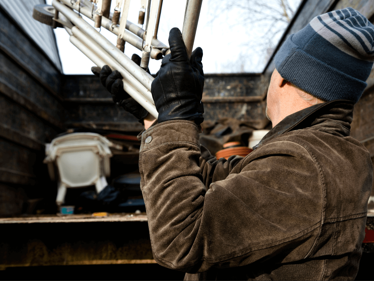 Man wearing a blue beanie, brown jacket, and black gloves lifting a folded metal chair into the back of a truck or dumpster containing assorted items.