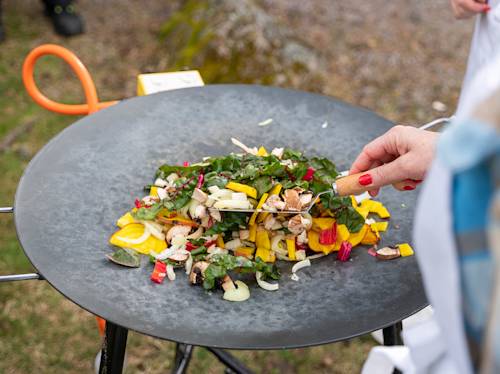 En person som lagar en färgglad grönsaksröra, inklusive paprika, svamp och grönsaker, på en rund metallgrill utomhus.