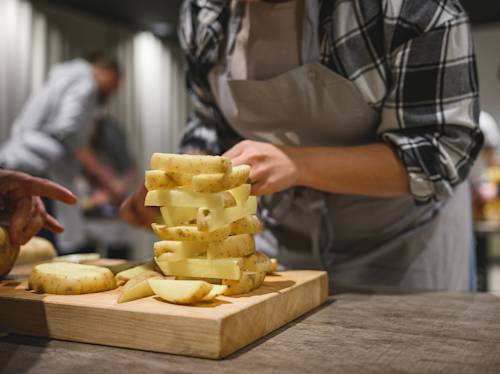 Person in an apron arranges thick-cut raw potato slices on a wooden cutting board; others are cooking in the background.