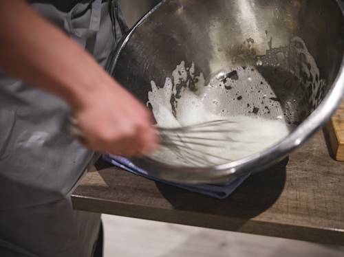 Person whisking egg whites or cream in a large metal mixing bowl on a wooden countertop, with some foam forming inside the bowl.