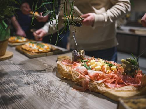 A charcuterie board with cheese, meats, and pickles sits on a wooden table. People are serving themselves food in the background.
