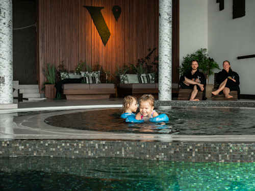 Två små barn leker i en rund inomhuspool medan två vuxna sitter på solstolar i bakgrunden.