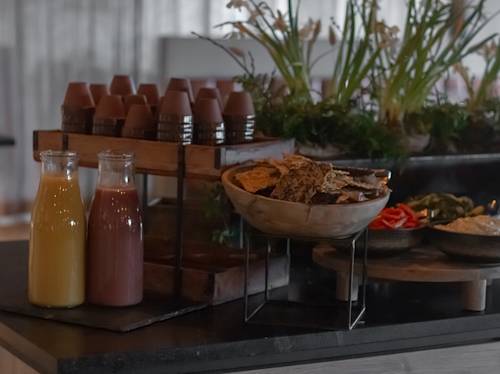 Glasflaskor med apelsin- och rödjuice, koppar staplade på en bricka och skålar med kex och grönsaker står uppställda på en bänkskiva med växter i bakgrunden.