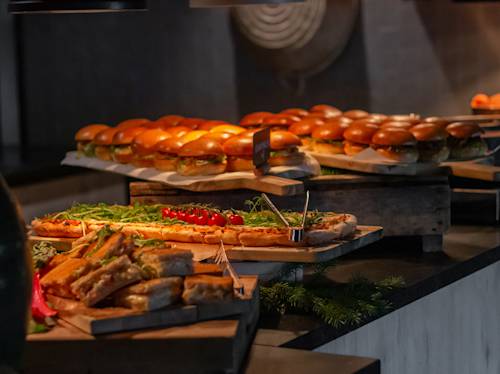 På ett buffébord står rader av hamburgare, tunnbrödspizzor toppade med grönsaker och tomater samt diverse smörgåsar på träskivor.