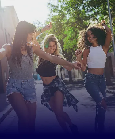 Three young women laughing and holding hands while walking down a sun