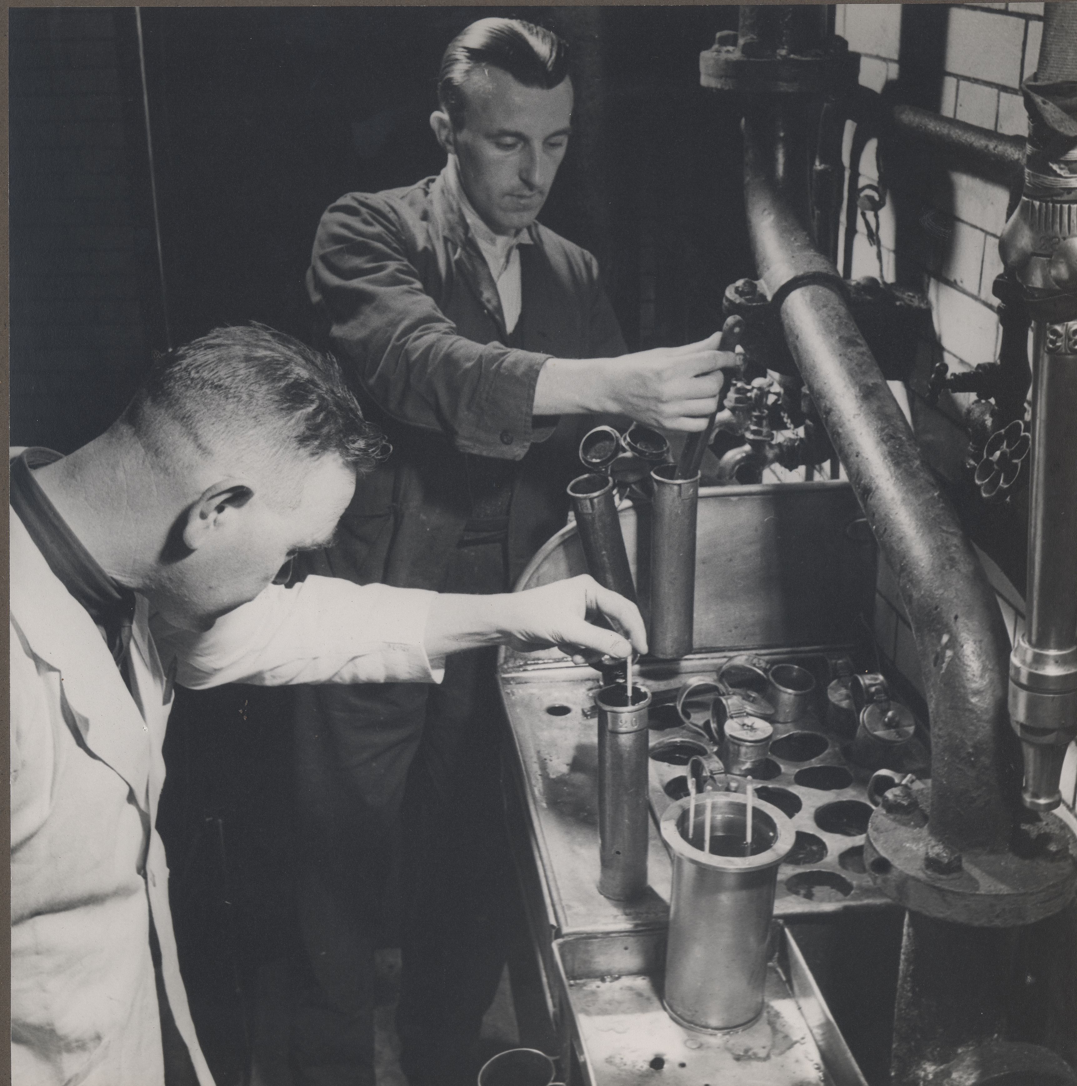 Vintage black-and-white photo of two brewery workers testing samples in metal cylinders beside large industrial pipes.