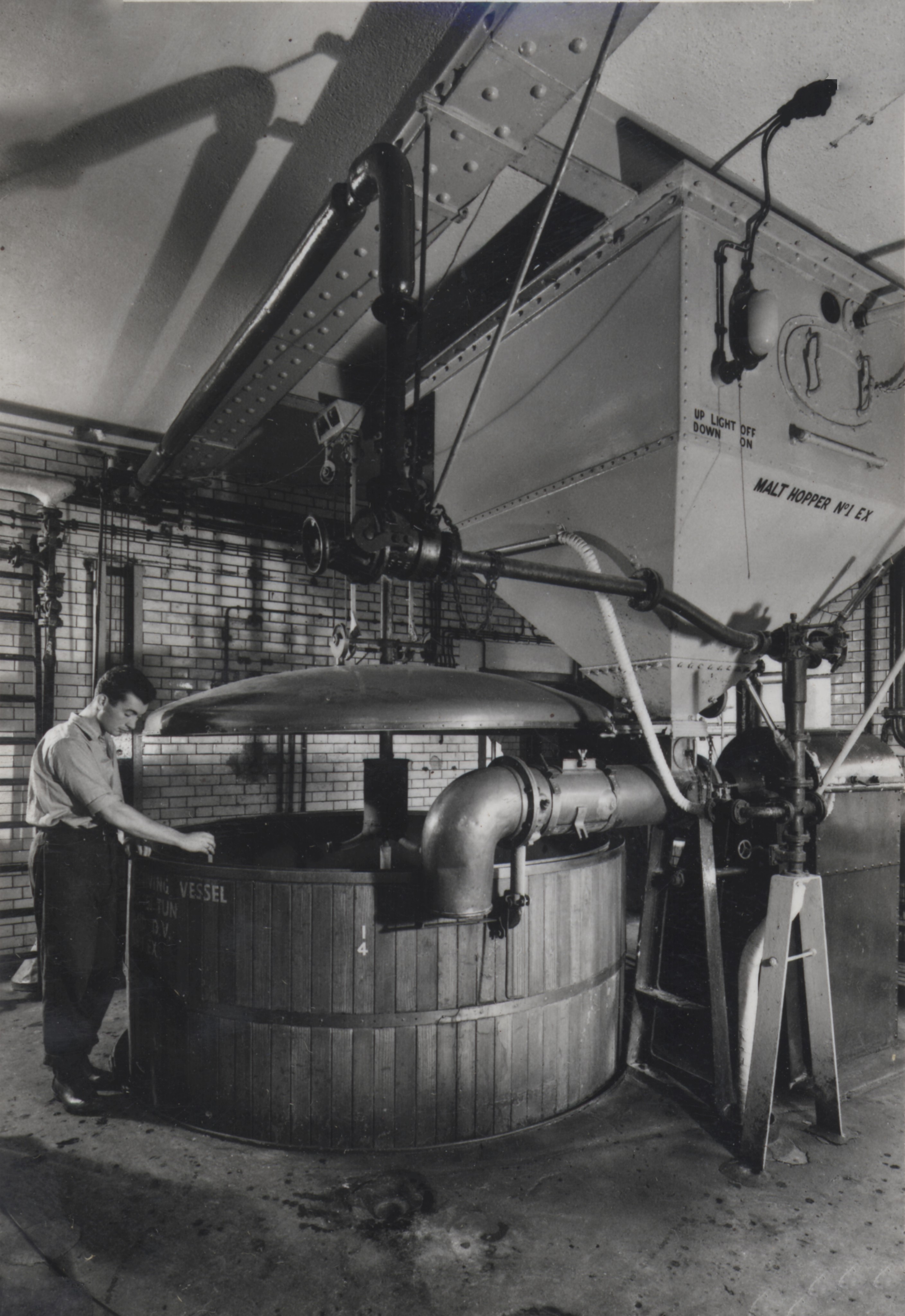 Vintage black-and-white photo of brewery worker standing beside large wooden mash tun under metal malt hopper and pipes.