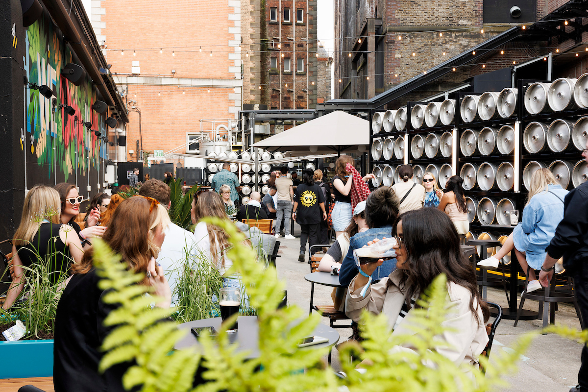 Crowded outdoor beer garden with people drinking at tables, a colorful mural, stacked metal kegs, and string lights overhead.