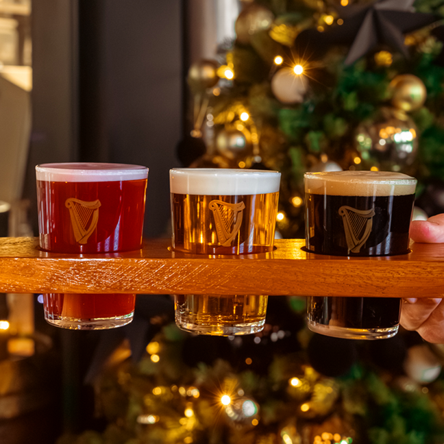 Hand holding a wooden beer flight with three small Guinness tasters in front of a blurred, glowing Christmas tree.