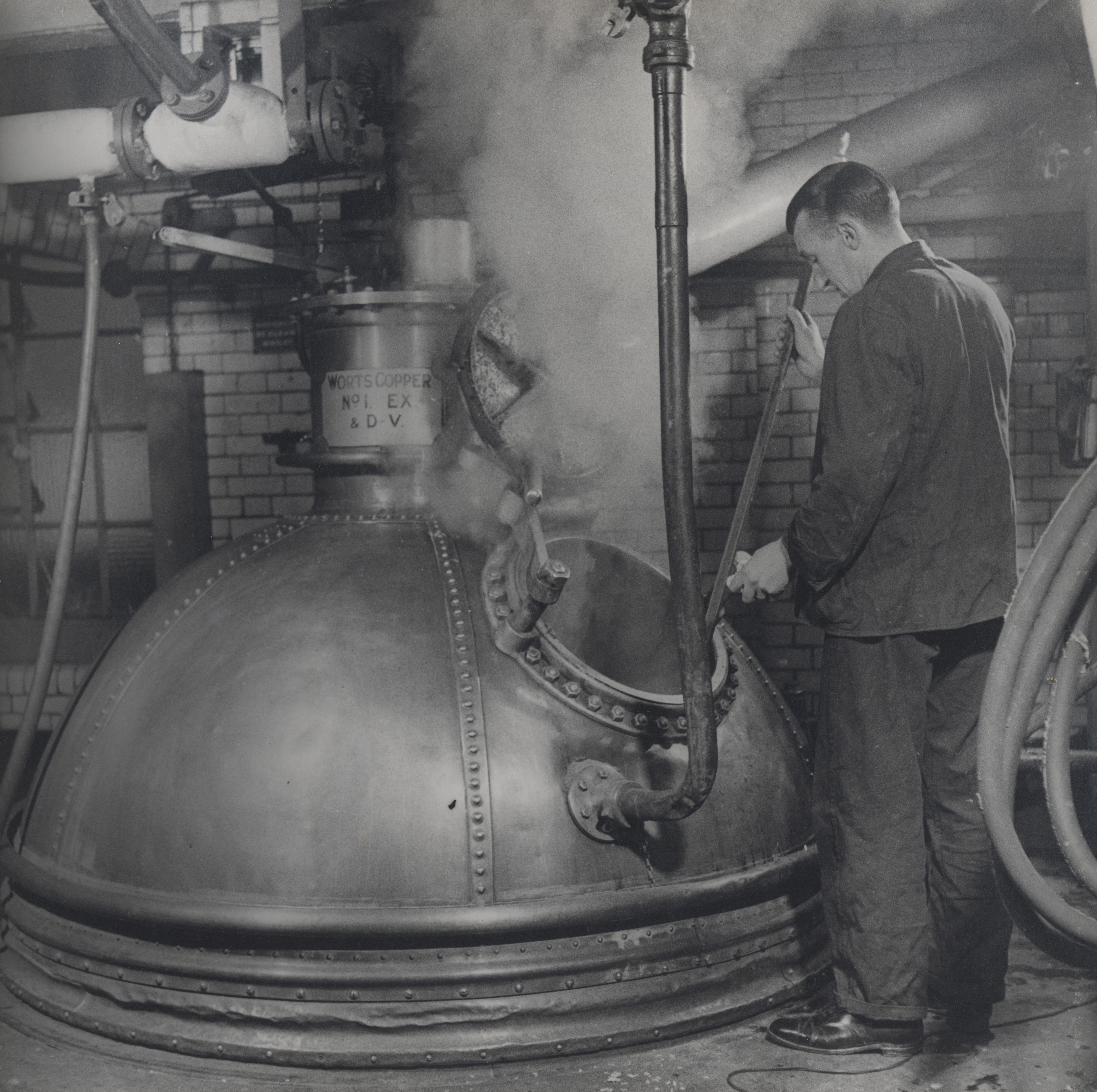 Vintage black-and-white photo of brewery worker stirring a steaming copper brewing vessel in an industrial room.