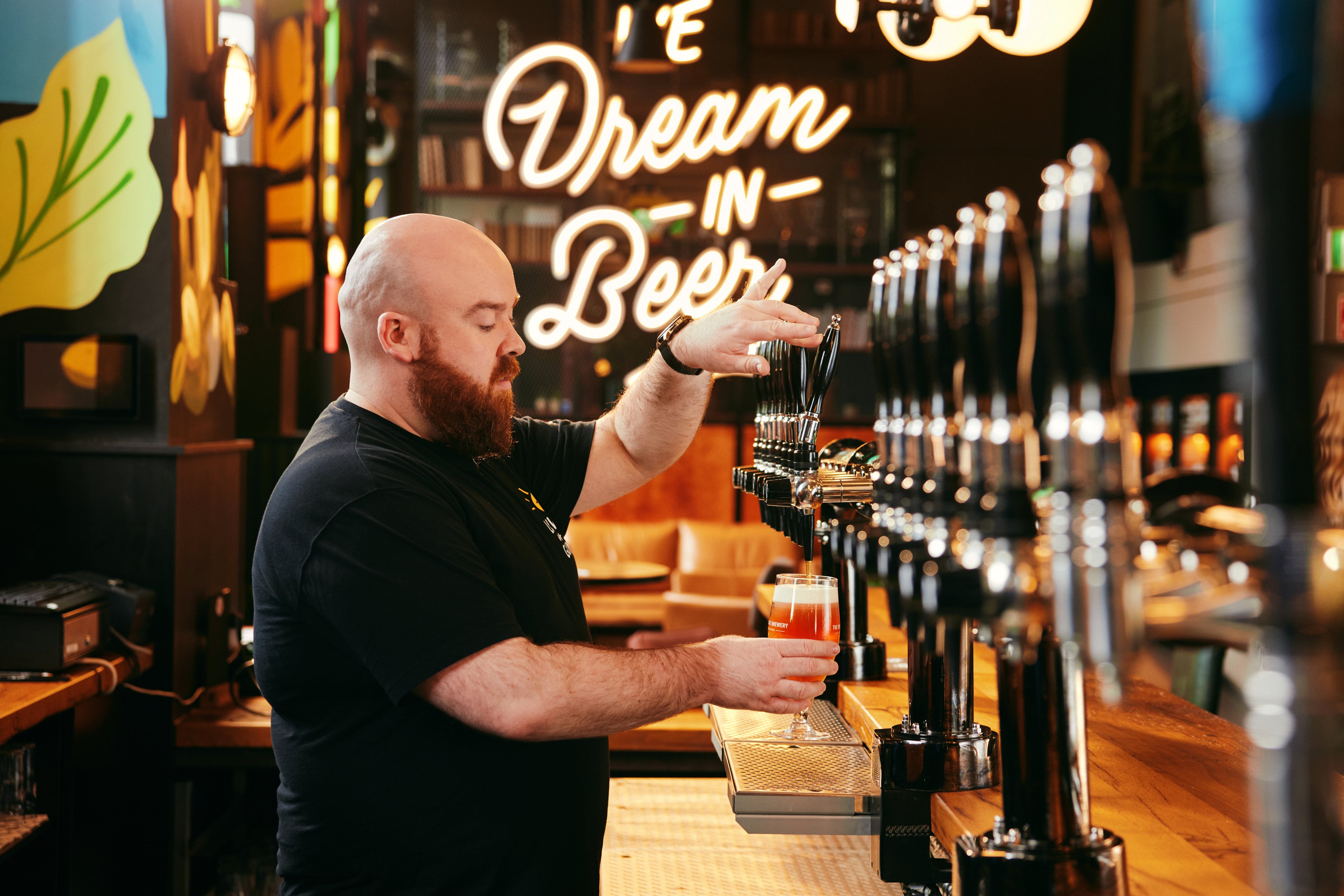 Bartender pours draft beer at a bar, standing by a row of taps with a glowing “Dream in Beer” sign in the background.