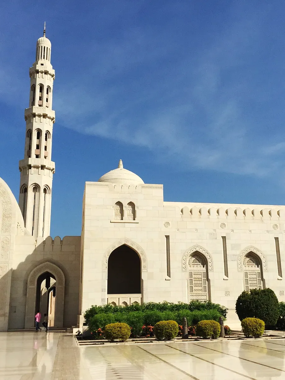 White mosque with tall minaret and dome against bright blue sky, landscaped with green shrubs and colorful flowers.