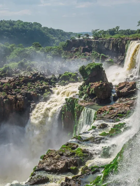 Powerful waterfall cascading over rocky cliffs surrounded by lush green forest, with mist rising from the rushing waters below.