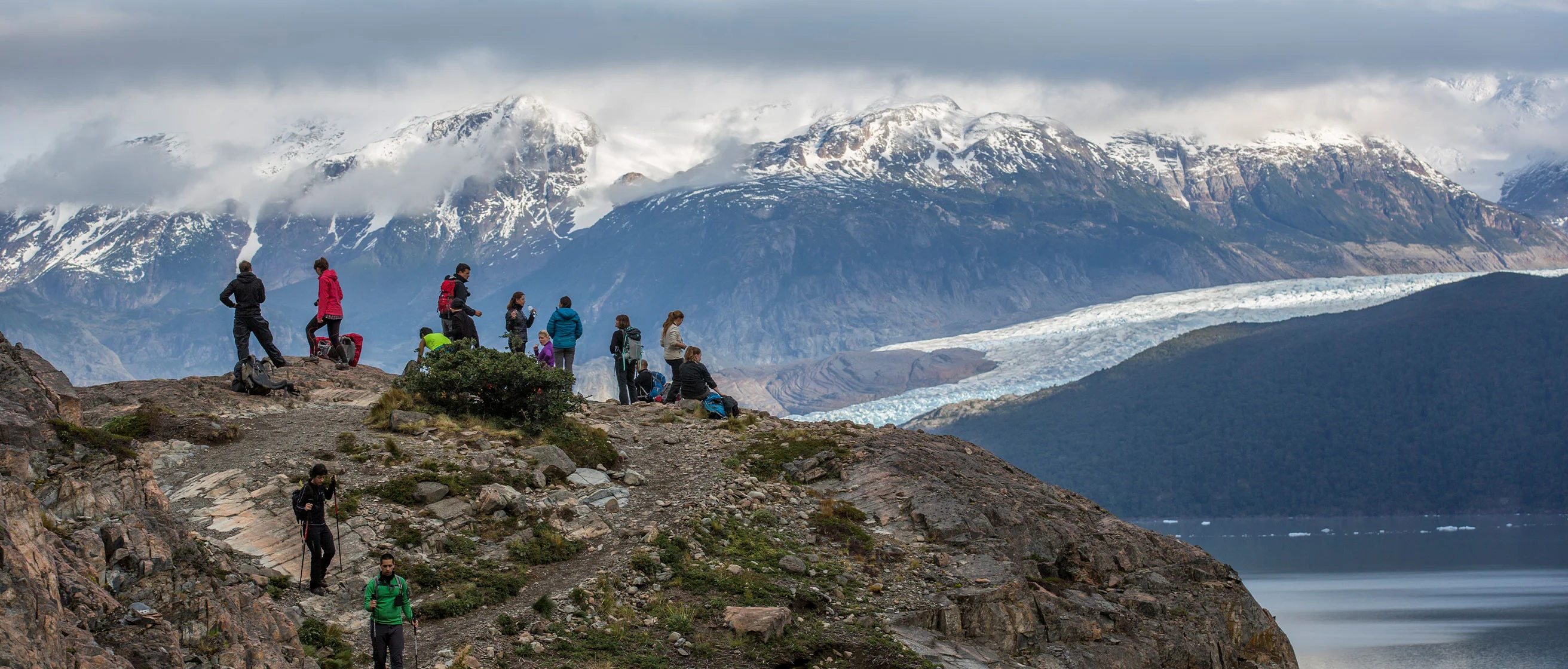 Hikers on rocky overlook admiring snow-capped mountains and glacier with cloudy sky in background.