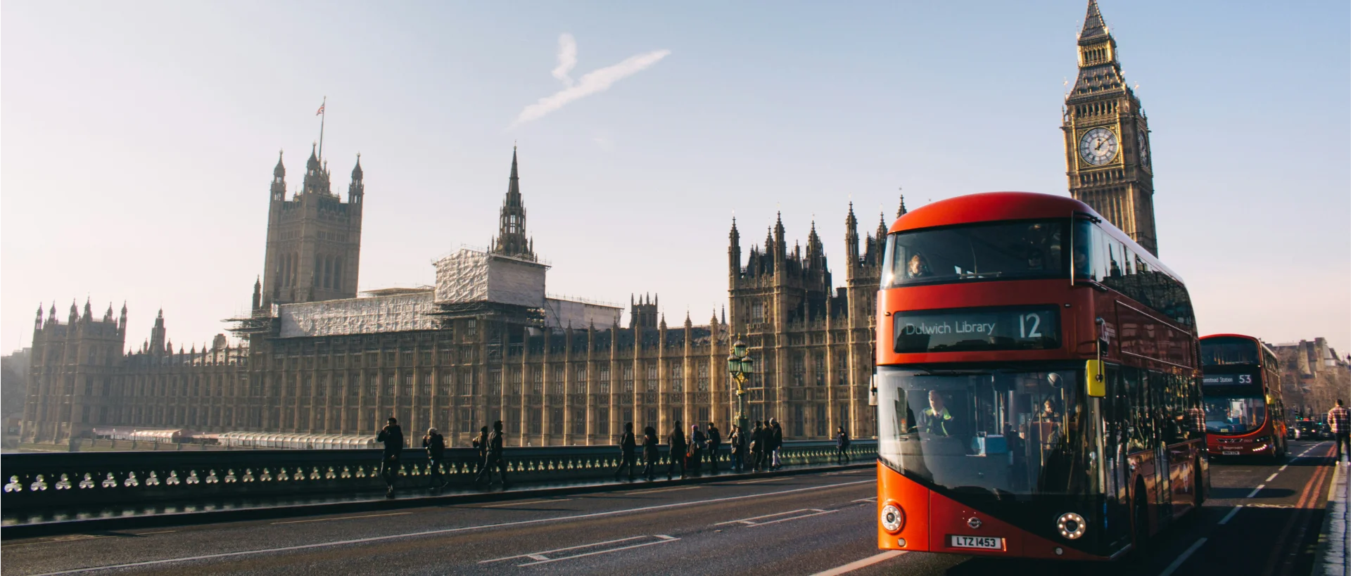 Red double-decker bus passing the Houses of Parliament and Big Ben in London under clear blue sky.