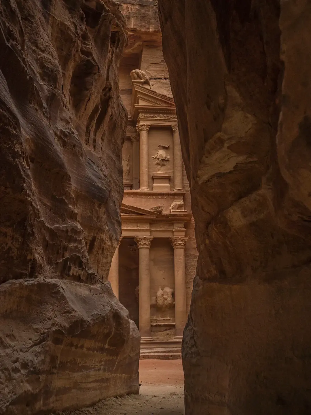 View of Petra's Treasury through the narrow Siq canyon, showing the ancient sandstone facade carved into the rock face.