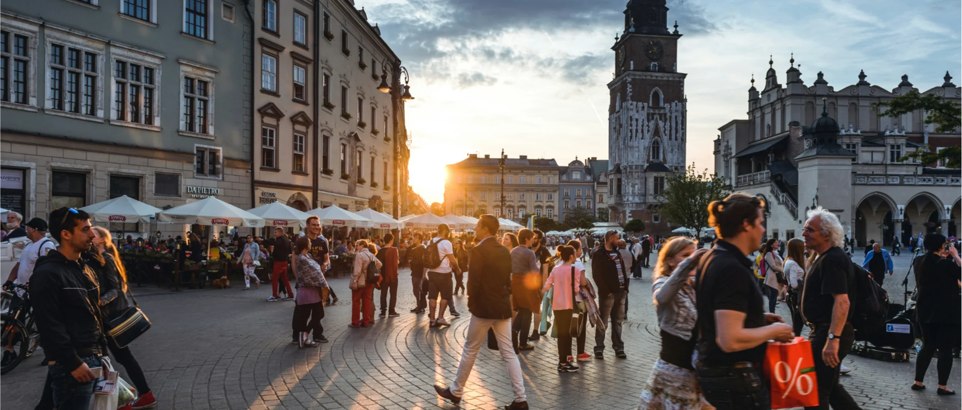 Sunset over Krakow's Main Square with crowds of people walking among outdoor cafés, historic buildings and a church tower.