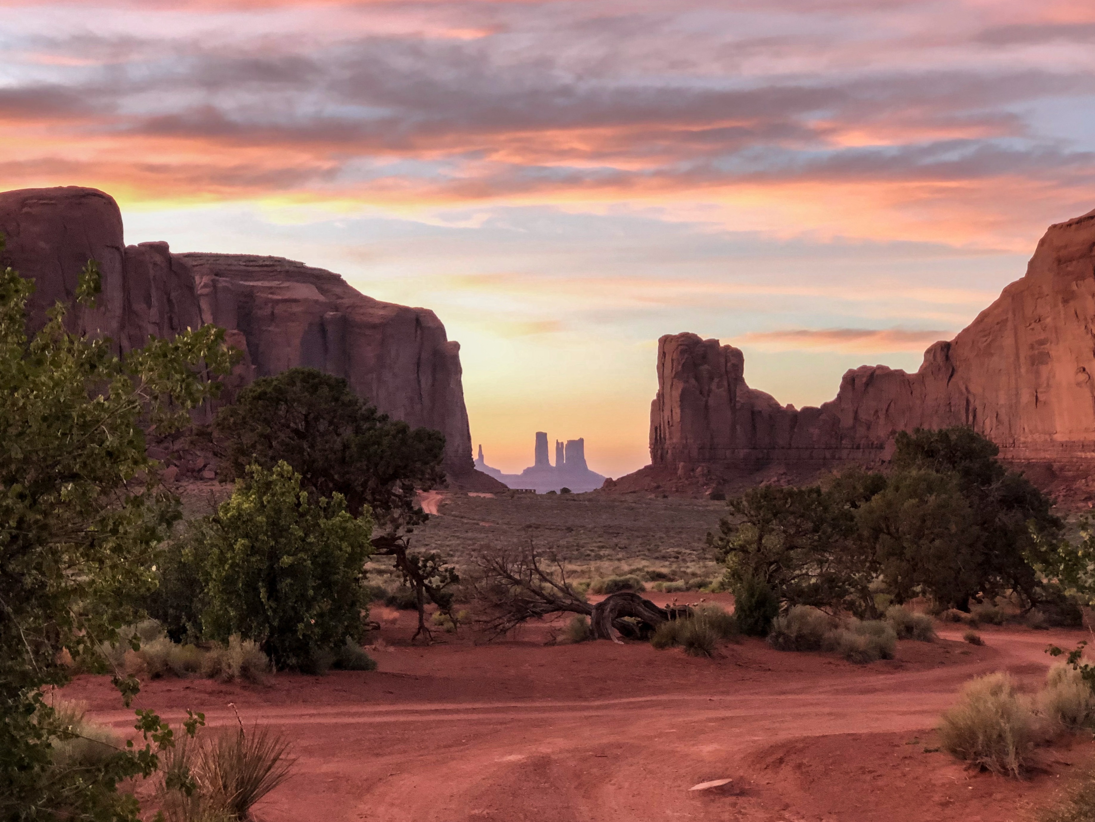 Sunset over Monument Valley with red sandstone formations silhouetted against a colorful sky, desert vegetation in foreground.