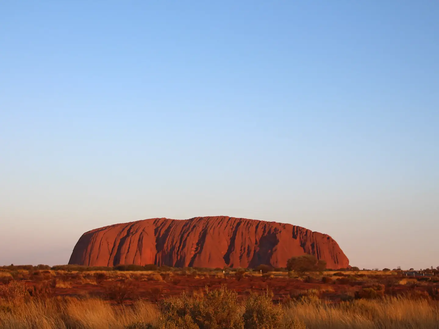 Uluru (Ayers Rock) glowing red at sunset against a clear blue sky, surrounded by arid Australian outback landscape.