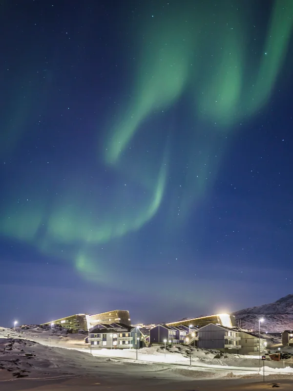 Green northern lights dance across a dark blue night sky above illuminated buildings in a snowy Arctic landscape.