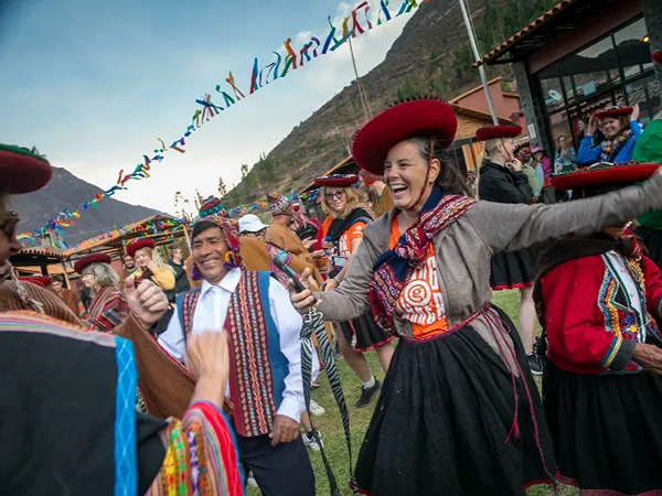People in colorful traditional Peruvian attire dance joyfully outdoors, with festive bunting overhead and mountains in the background.