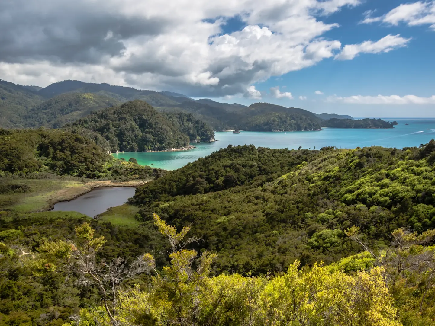 Panoramic view of lush green hills and mountains surrounding turquoise bay waters under a partly cloudy blue sky.