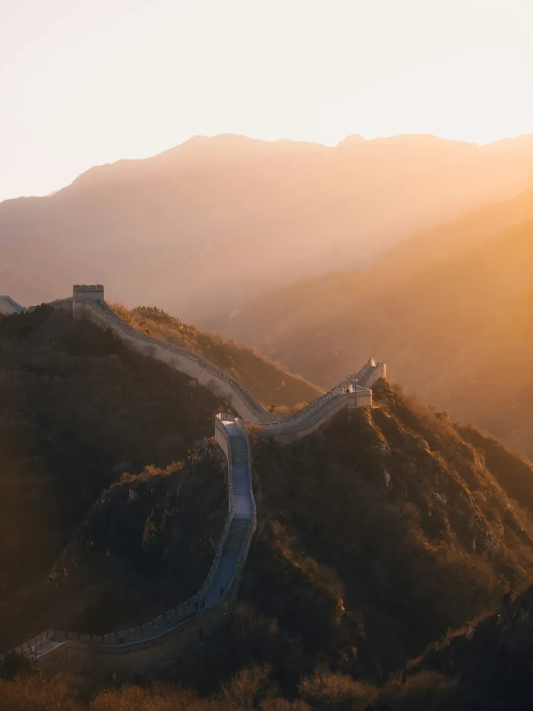The Great Wall of China winding along mountain ridges at sunset, bathed in golden light and long shadows.