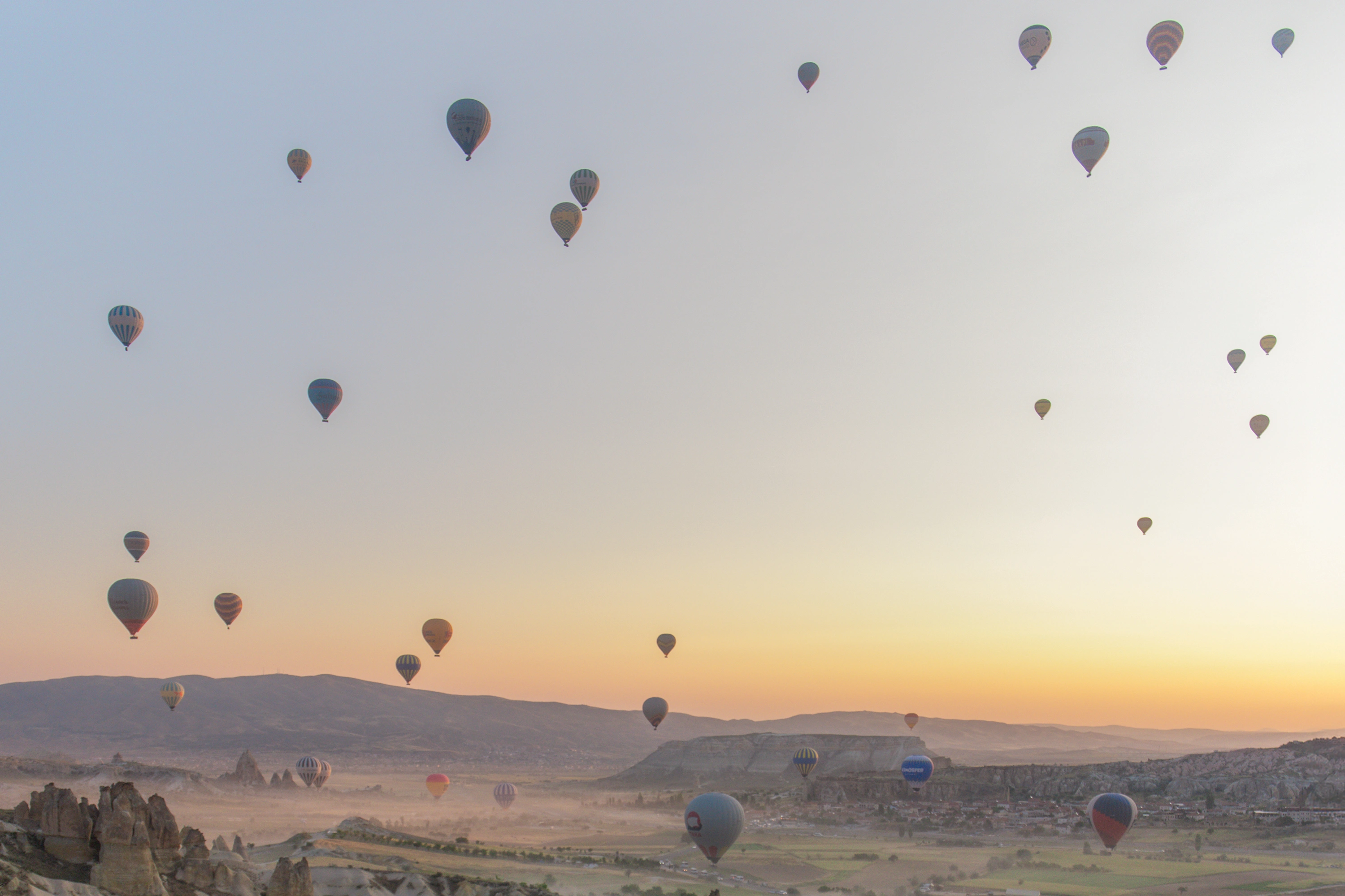 Dozens of colorful hot air balloons float over Cappadocia's rocky landscape at sunrise, with a warm golden glow on the horizon.