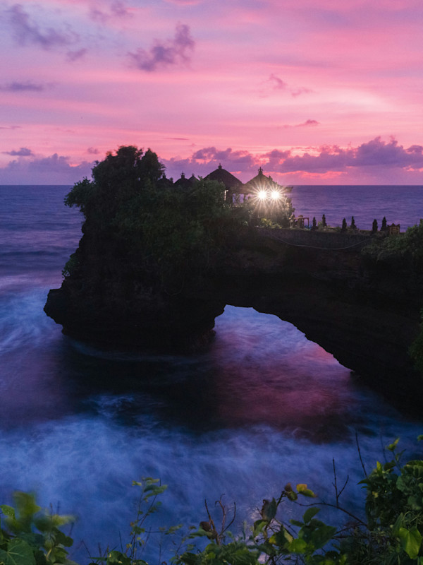 Tanah Lot temple perched on a rocky outcrop against a vibrant purple-pink sunset sky, with ocean waves crashing below.