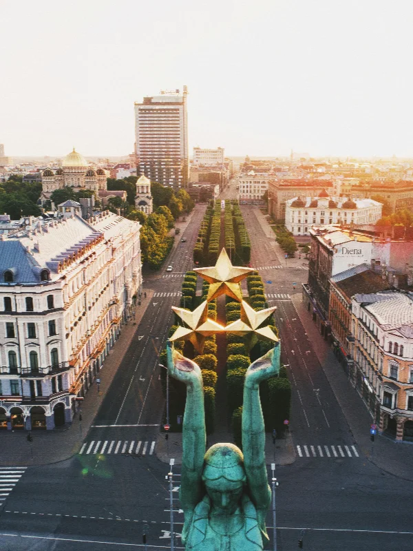 Aerial view of a European city boulevard with a green-patina statue holding golden stars, historic buildings, and a tall skyscraper.