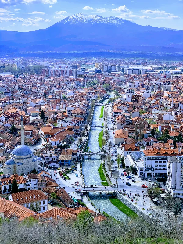 Aerial view of Prizren, Kosovo with a river flowing through the city center, surrounded by buildings and snow-capped mountains in the distance.