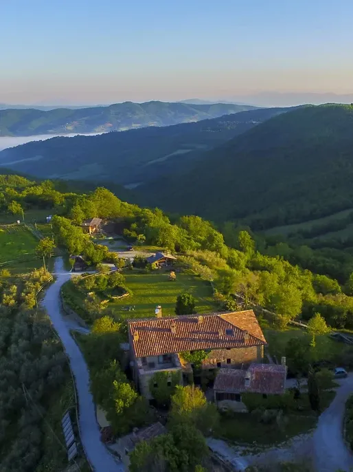 Aerial view of a rustic stone house with terracotta roof nestled in green hills, with misty mountain ranges stretching to the horizon.