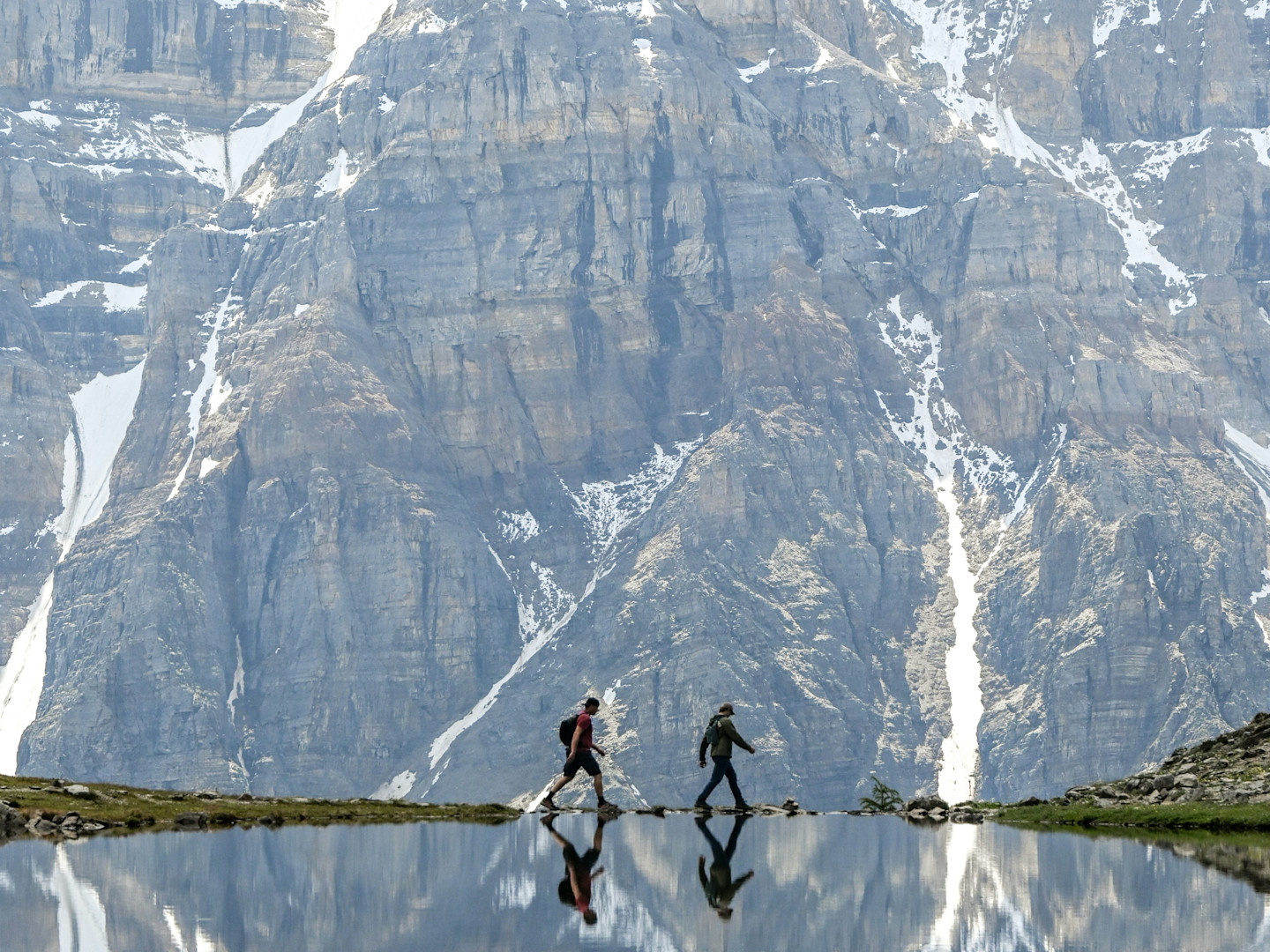 Two hikers walking along a mountain lake with massive snow-streaked rocky cliffs towering in the background.
