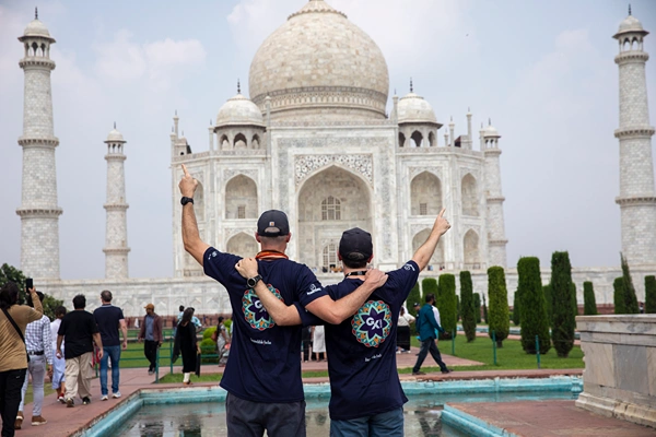 Two men in navy blue shirts pose with arms raised in front of the iconic white Taj Mahal in Agra, India.