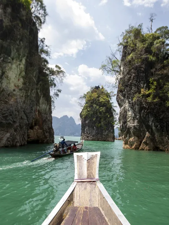 Wooden boat navigating through turquoise waters between towering limestone cliffs, with another tour boat visible ahead.