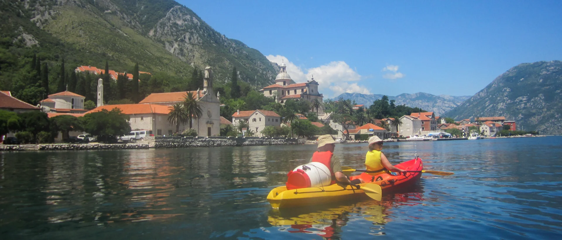 Kayakers in yellow and red boats paddling on blue water near a coastal Mediterranean village with orange-roofed buildings and mountains.