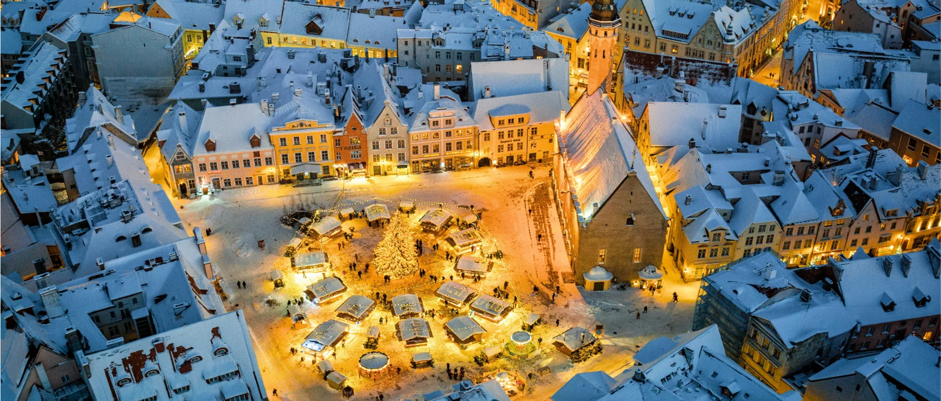 Aerial view of a snowy European town square with Christmas market stalls, illuminated buildings, and snow-covered rooftops at night.