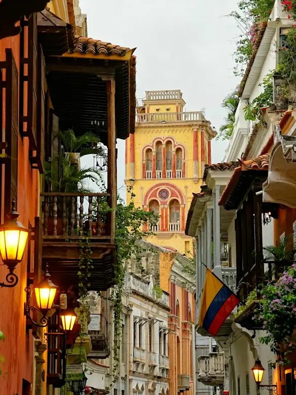 Narrow street in Cartagena with colorful colonial buildings, glowing lanterns, a Colombian flag, and flowering balconies.