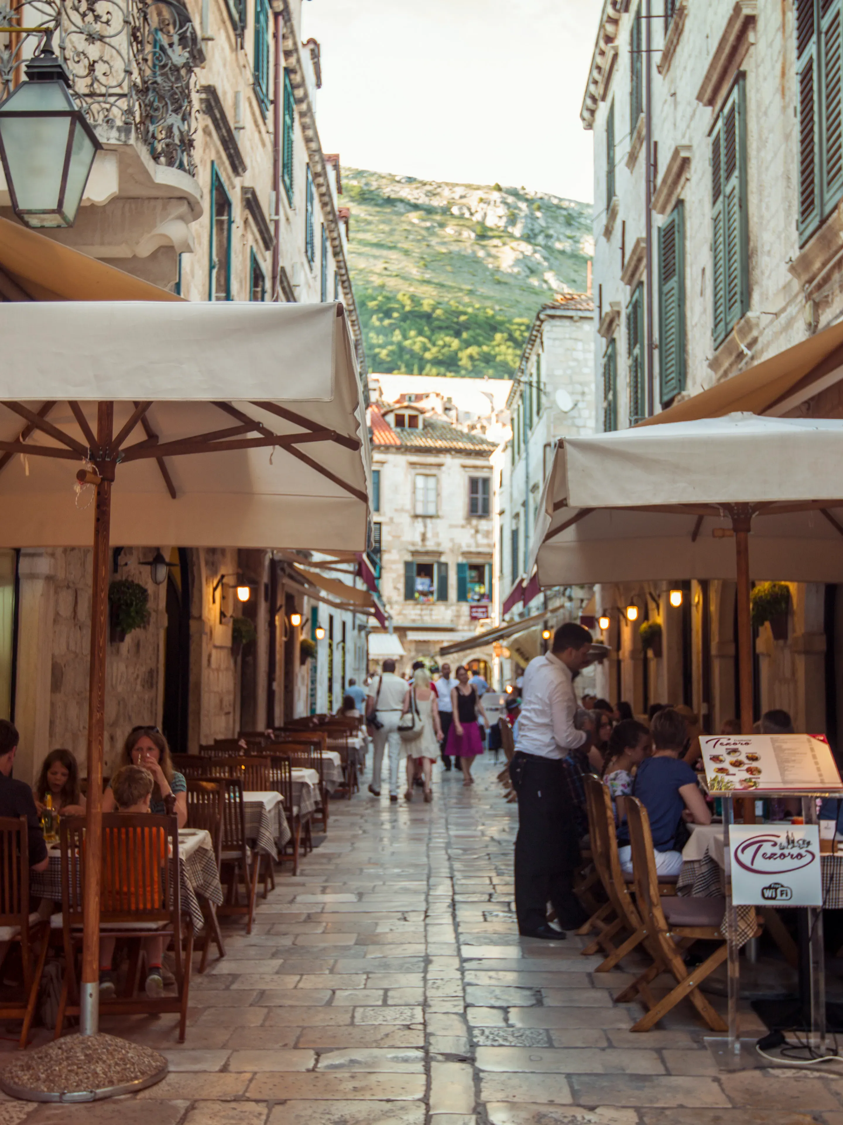 Narrow cobblestone street in Mediterranean town with outdoor cafes, umbrellas, and diners beneath historic buildings and mountain views.